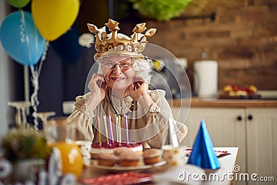 Cheerful lovely senior woman wearing inflatable golden crown and feeling joyful sitting at the table celebrating birthday Stock Photo