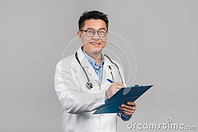 Cheerful mature korean man doctor in white coat, glasses with stethoscope writes at tablet Stock Photo