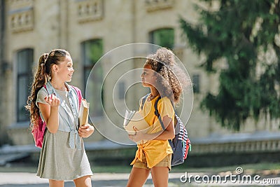 Cheerful multicultural schoolgirls holding books and talking in schoolyard Stock Photo