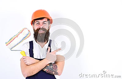 Cheerful painter, decorator on smiling face holds paint roller, white background, copy space. Man with beard in helmet Stock Photo