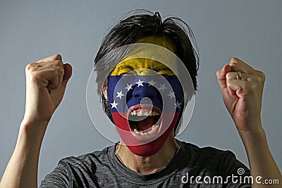 Cheerful portrait of a man with the flag of Venezuela painted on his face on grey background. The concept of sport or nationalism. Stock Photo