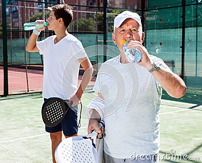 Mature man and young man drink water on padel court Stock Photo