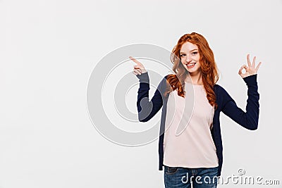 Cheerful pretty young redhead lady showing okay gesture while pointing. Stock Photo