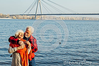 Cheerful senior citizens woman and man are standing and hugging on the lake, against the background of the bridge Stock Photo