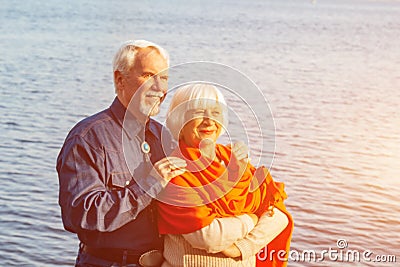 Cheerful senior citizens woman and man are standing and hugging on the lake, against the background of the bridge Stock Photo
