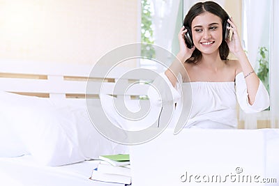 Cheerful smile woman listening music with headphones, book and laptop feeling relax sitting on white bed in the morning. Stock Photo