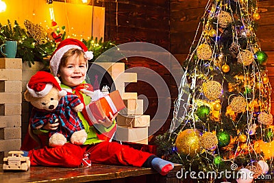 Cheerful smiling baby Santa boy holding his teddy bear and Christmas present near fire place and decorated New Year tree Stock Photo