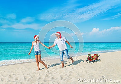 Cheerful smiling young couple in red Santa hats walking at tropical ocean sandy beach with sleds decorated fir-tree and golden fan Stock Photo