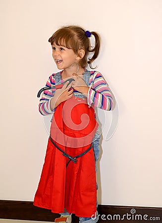 The cheerful three-year-old girl tries on on herself a beautifulred dress on a hanger Stock Photo