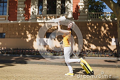 Cheerful traveler tourist woman in yellow summer casual clothes, hat with suitcase city map walk in city outdoor. Girl Stock Photo