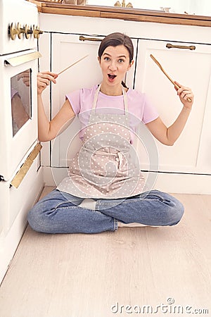 Cheerful woman in apron sitting on kitchen floor holding cooking utensils Stock Photo