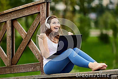 Cheerful woman in wireless headphones makes telework with laptop outdoors. Stock Photo