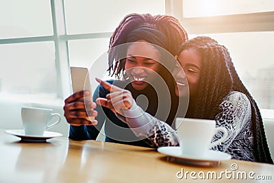 Cheerful young african women sit together. They smile and point on white phone. Models sit ar bringht window. Stock Photo