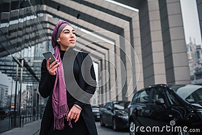 Cheerful young arabian model stand outside and pose. Woman hold phone and look to right. She stands close to black cars. Stock Photo