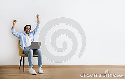 Cheerful young arabic guy in casual and glasses sits on armchair with laptop, makes gesture of victory Stock Photo