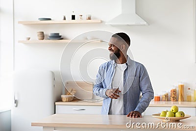 Cheerful young bearded black guy with glass of clean water in hand in kitchen interior looks at window Stock Photo