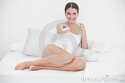 Cheerful young brown haired model in white pajamas watching tv while eating popcorn Stock Photo