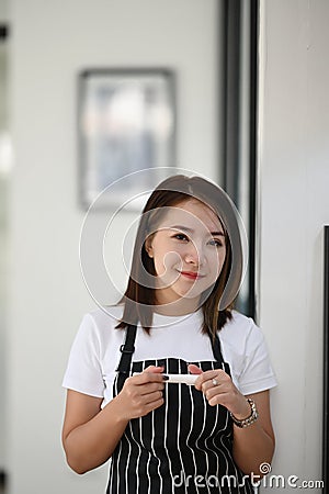 A young female entrepreneur wearing black apron standing in her own modern coffee shop. Stock Photo