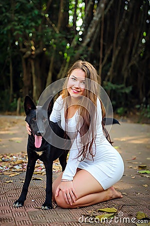 Cheerful young girl on walk in the park with her four-legged friend. Pretty woman in short dress and black dog playing outdoors. Stock Photo