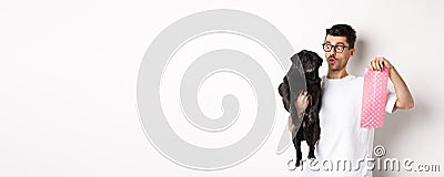 Cheerful young man holding black pug and pink dog poop bag, standing over white background Stock Photo