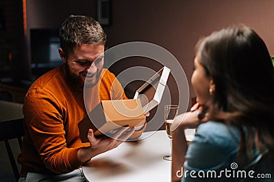 Cheerful young man opening box with received gift from loving girlfriend enjoying romantic dinner date on birthday or Stock Photo