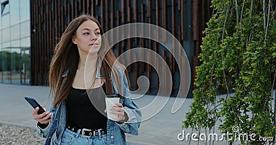 Cheerful young woman is walking on the street and drinks coffee and using smartphone. Woman messaging on phone at street Stock Photo