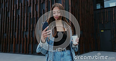 Cheerful young woman is walking on the street and drinks coffee and using smartphone. Woman messaging on phone at street Stock Photo