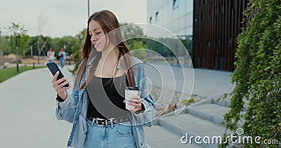 Cheerful young woman is walking on the street and drinks coffee and using smartphone. Woman messaging on phone at street Stock Photo