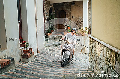 Cheerfully smiling man in helmet and sunglasses riding the motor scooter on the narrow Sicilian old town streets. Happy Italian Stock Photo