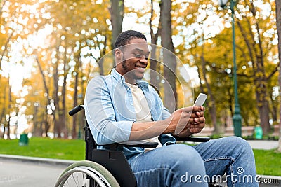 Cheery disabled black guy in wheelchair using mobile phone, checking email or messages at autumn park Stock Photo