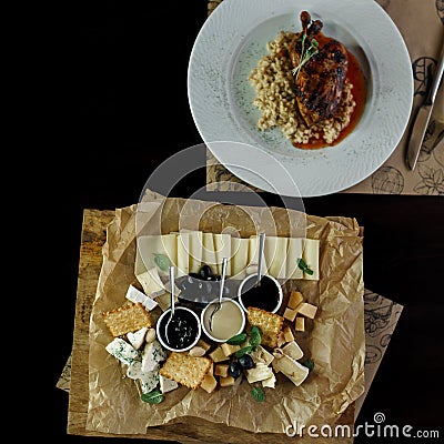 Cheese platter stands on a wooden table in a restaurant next to a duck leg in a sauce. Easy tasty snack. Useful lunch in a cafe. Stock Photo