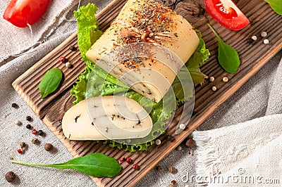 Cheese roll with olive herbs on a lettuce sheet. A piece is cut off. Cheese on a wooden board. Decorated with arugula, spices and Stock Photo