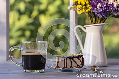 Cheesecake with chocolate sauce and glass cup of black coffee on a wooden table near window. Coffee time with slice of cake at Stock Photo
