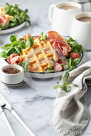 Cheesy belgian waffles served with ham, tomatoes and lettuce corn with cutlery on white marble background . Savory Stock Photo