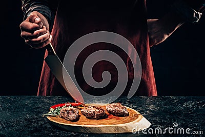 Chef in apron with meat fork and knife slicing gourmet grilled steaks with rosemary and chili pepper on wooden board Stock Photo