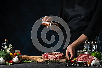 The chef in black apron pours pepper on fresh meat steak on wooden chopping board at the professional restaurant or hotel kitchen Stock Photo