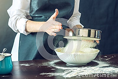 The chef in black apron sifts the flour through a sieve to prepare the dough for pizza Stock Photo