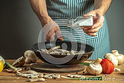 A chef in a blue apron is sprinkling salt on mushrooms in a frying pan. Concept of process cooking food Stock Photo