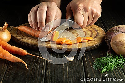 Chef cuts fresh carot on a wooden cutting board. Close-up of cook hands while preparing vegetarian food. Copy space Stock Photo