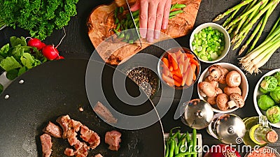 Chef cuts green onions while frying fresh meat in a wok, on a background with vegetables Stock Photo