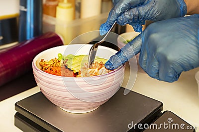 Chef's hands gloved in plastic preparing a salmon poke bowl with masago roe and avocado Stock Photo