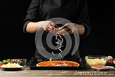 The chef sprinkles pizza with mazzarella cheese. The concept of delicious food and healthy food. On a black background for design Stock Photo