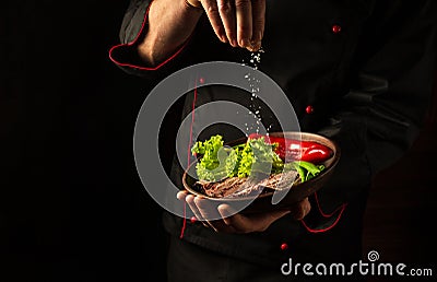 The chef sprinkles salt on a sliced steak with beef and vegetables in a plate. The concept of serving dishes to order Stock Photo