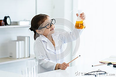 In chemistry classroom with many laboratory tools on shelves and table. A little Asian girl with glasses looking yellow chemical Stock Photo