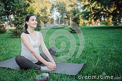 Cherful nice young woman stretching legs on yoga mate outside in park. She smiles and hold hands on feet. Stock Photo