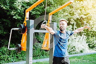 Chest workout with band crossovers. Charismatic man doing exercise on the outdoor exercise machine. Stock Photo