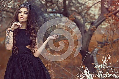 Chic young woman with perfect make up wearing lace dress and black jewel crown with veil standing in the abandoned garden Stock Photo