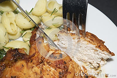 Chicken breast fried with spices, boiled pasta with herbs, metal fork and knife which cut a piece of the breast on a white plate Stock Photo