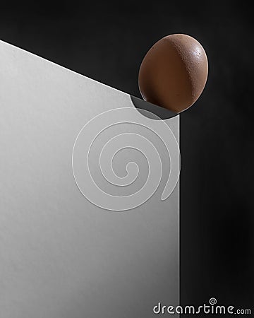 A chicken egg is precariously balanced on the edge of a piece of furniture on a black background Stock Photo