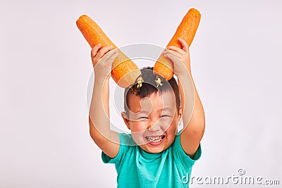 Child boy in turquoise shirt, holds huge carrots depicting horns - fruits and healthy food Stock Photo